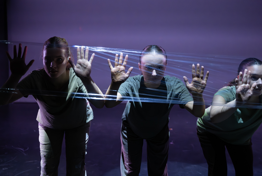 Three dancers leaning towards the camera with their hands pressed against cellophane.