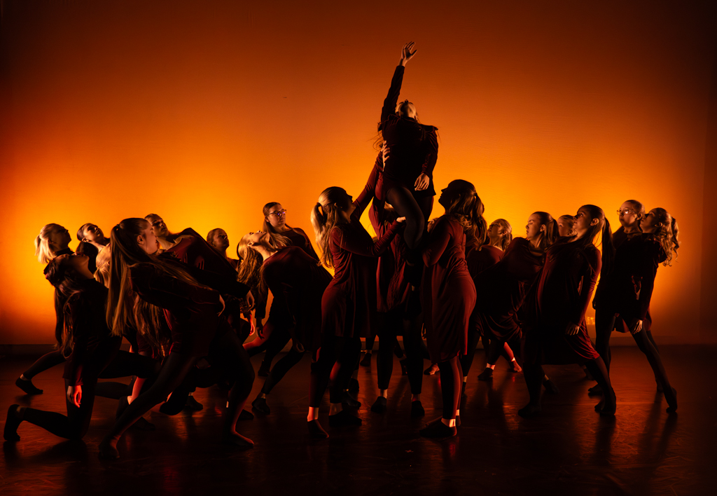 Group of dancers gathered in a circle in the centre of the stage all gazing up at one of them being held up with their hand stretched upwards