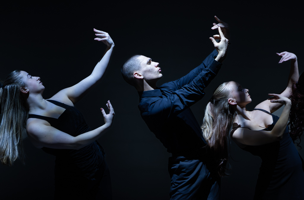 Three dancers in dark clothing all stood with their hands up in a curved position as they look up 