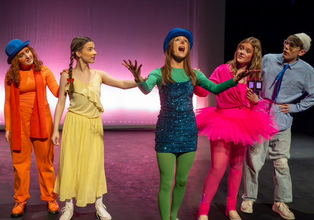 Five students all in a line, looking at the student in the middle as she sings. All of them are wearing fancy dress costumes. 