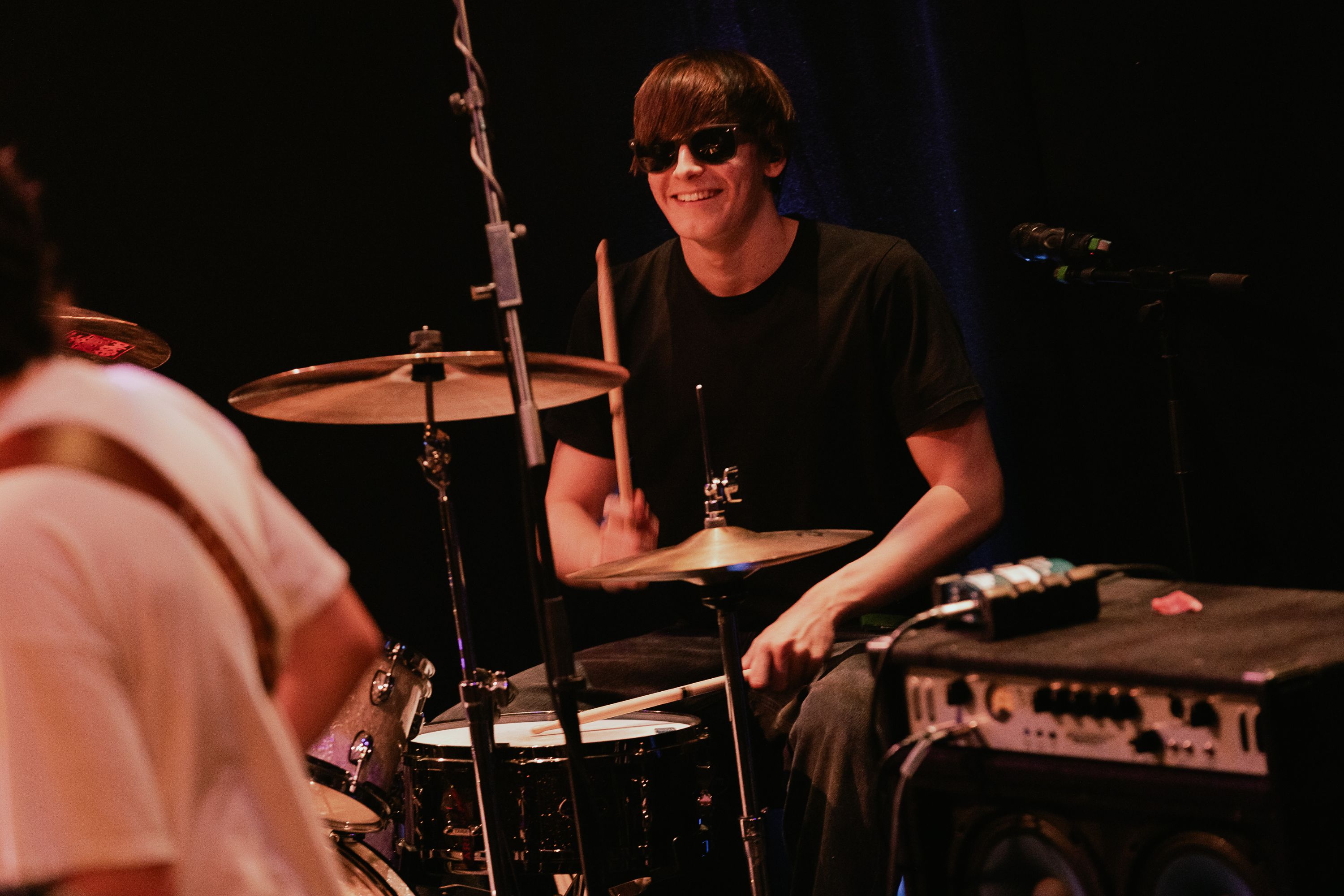 A student wearing sunglasses smiling whilst playing the drums. 