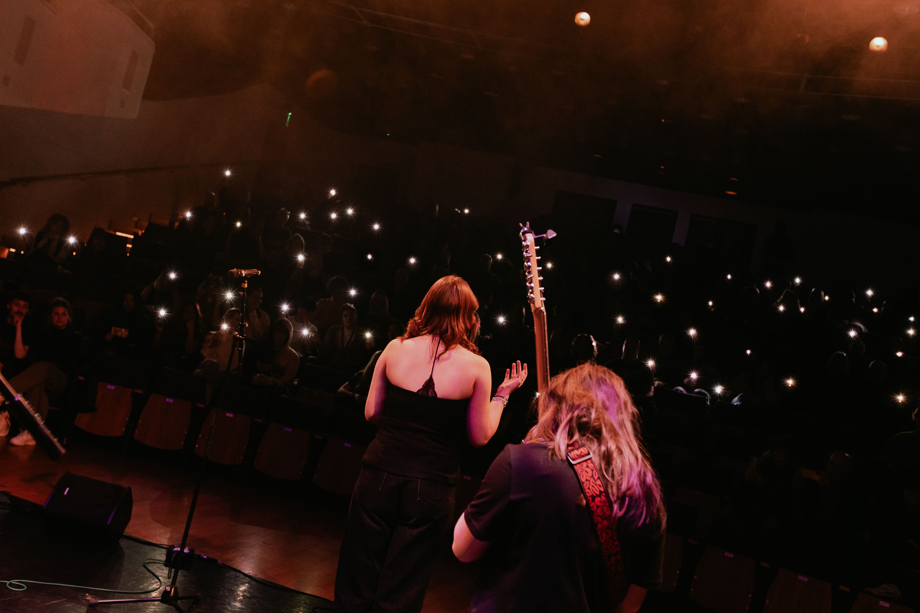 A band performs on a stage in front of an audience with their phone torches turned on to create a starlight effect in the darkness of the audience.