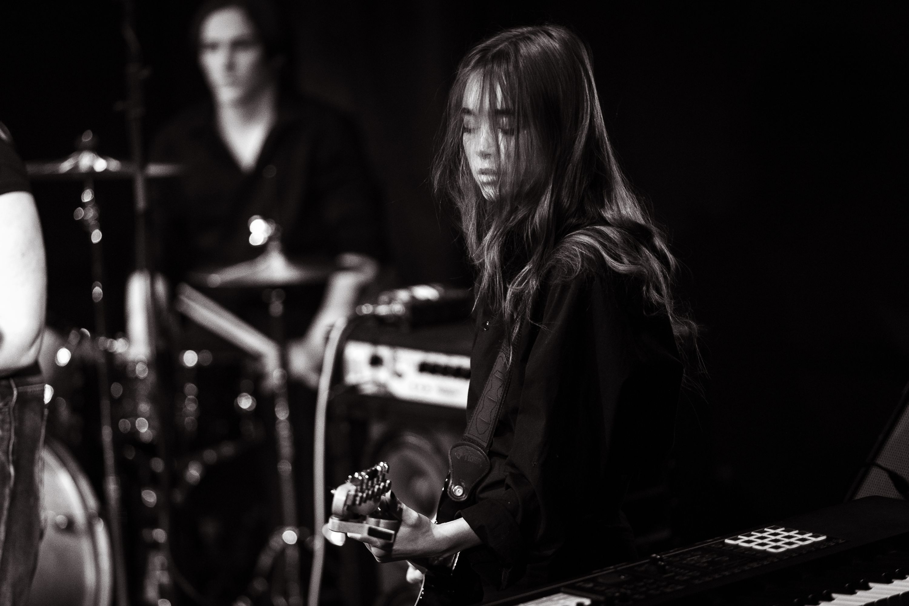 A student playing the guitar with a drummer in the background.